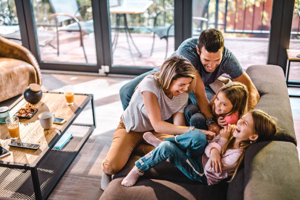 Above view of playful parents tickling their daughters at home. High angle view of cheerful parents having fun while tickling their daughters on sofa in the living room. lifestyle stock pictures, royalty-free photos & images