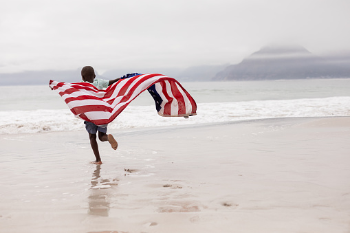 Rear view of African american boy running with American flag on the beach