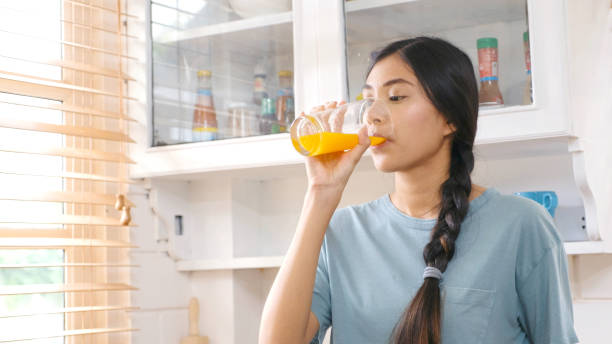 istock Young beautiful asian woman drinking orange juice while standing by window in kitchen background, peolpe and healthy lifestyles 1159551977