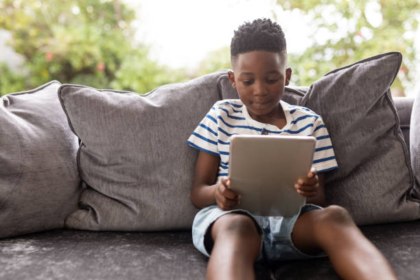 menino que usa a tabuleta digital em um sofá na sala de visitas - africa child reading african descent - fotografias e filmes do acervo