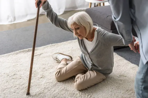 cropped view of man helping sick senior mother with cane fallen on floor