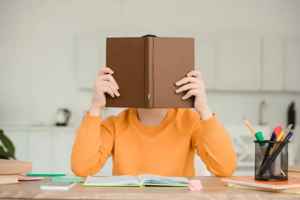 boy hiding face behind book while sitting at desk and doing schoolwork at home