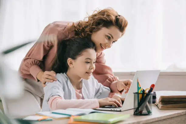 happy mother helping smiling daughter doing schoolwork at home