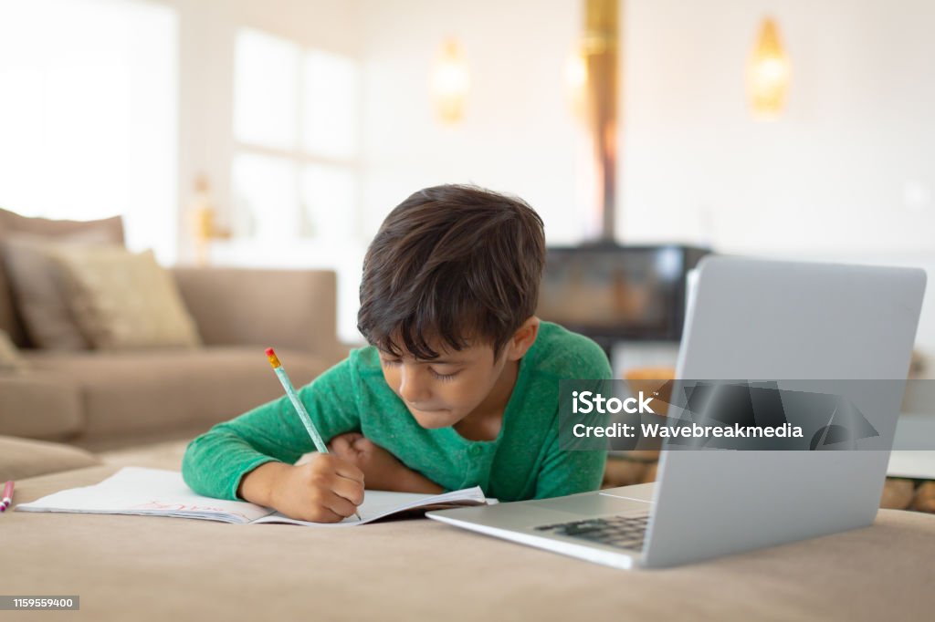 Boy using laptop while drawing a sketch on book at home Front view of African american boy using laptop while drawing a sketch on book at home Child Stock Photo