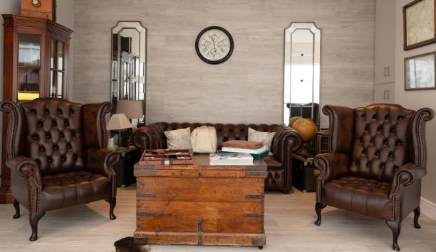 Digitally rendered inviting retro-style living room glowing under soft evening light. The space highlights mid-century modern armchairs, rich wood tones, classic wall sconces, and a tea set arranged on a round table. Ambient lighting from floor and wall lamps creates a relaxing mood, emphasizing the textured ochre walls, elegant curtains, and timeless floral artwork. Perfect for themes of home warmth, retro ambiance, and interior elegance.