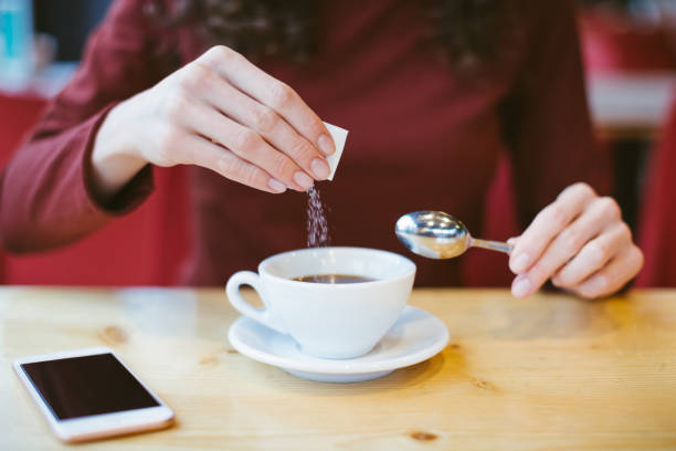 Woman's hands pouring sugar into black coffee - girl sitting at the table with espresso and smartphone - blood and glycemic index control for diabetes -excess of white sugar in food concept stock photo