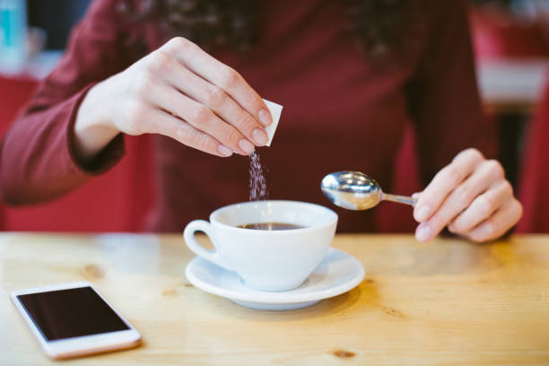 mãos da mulher que derramam o açúcar no café-menina preta que senta-se na tabela com café e smartphone-controle do índice do sangue e do glicêmico para o diabetes-excesso do açúcar branco no conceito do alimento - fat cell - fotografias e filmes do acervo