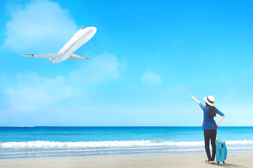 Rear view of asian woman in hat with suitcase bag standing on the beach and pointing to a flying plane above the sea