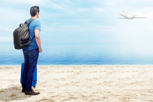 Rear view of asian man with suitcase bag and backpack standing on the beach with flying airplane above the sea and blue sky background