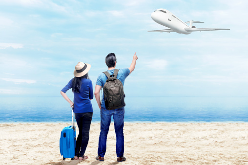 Rear view of asian couple with suitcase bag and backpack standing on the beach and pointing to a flying plane above the sea with blue sky background