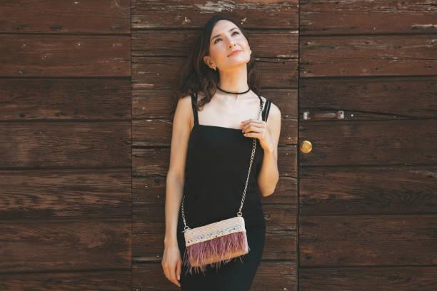 Beautiful woman with little crossbody bag looking up in the sky leaning on the old vintage wooden door stock photo