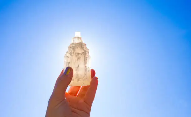 Person holding naturally carved mineral stone Selenite tower against sun and blue sky, stone has healing and cleansing properties. Also used for recharging other mineral crystal stones.