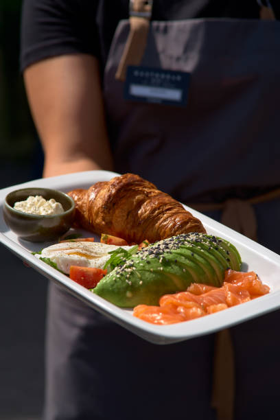 istock Breakfast is set. Whole grain sandwich with fried egg, red fish, avocado. Plate in the hands of a waitress serving breakfast in a restaurant. 1159578802
