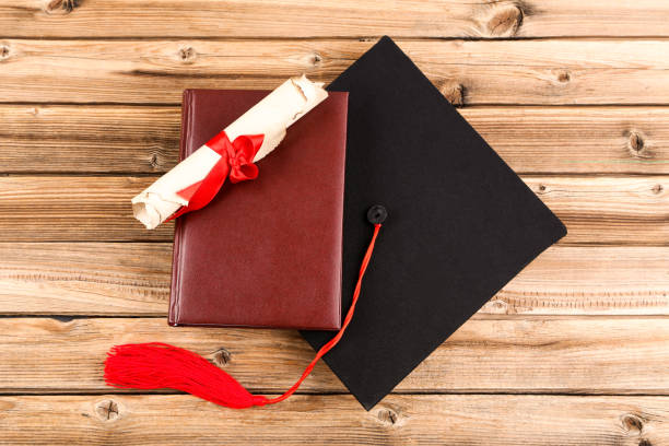 istock Graduation cap with diploma and book on brown wooden table 1159582707