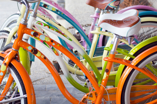 Row of colorful retro replica bikes. Each has a colorful dot design on the from and on the top of the seat. The first one is orange, second is white, then cream, blue, and lavender. The bikes are cropped out around the edges. The background is a light concrete and you cannot see much. 