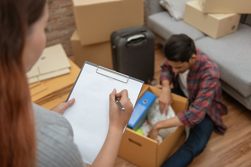 Young Asian couple packing their belonging into cardboard box by using a check list before moving to new resident or house after buy or rent a new one.