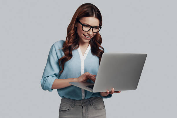 istock Very busy. Attractive young woman using laptop and smiling while standing against grey background 1159586890