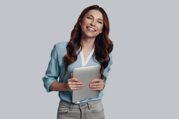 istock Enjoying her new tablet. Attractive young woman looking at camera and smiling while standing against grey background 1159587185