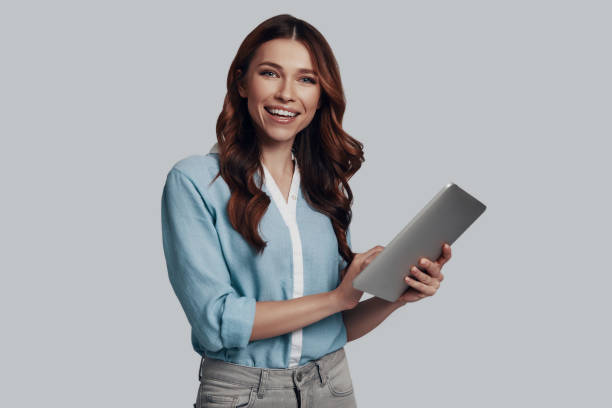 istock Examining her new tablet. Attractive young woman working using digital tablet and smiling while standing against grey background 1159587448
