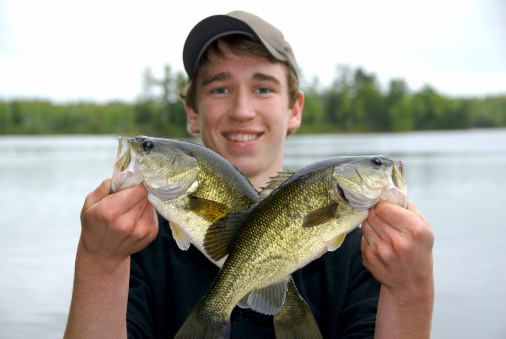Boy holding two largemouth bass in an x formation with a big smile and a baseball hat with a lake and trees in the background