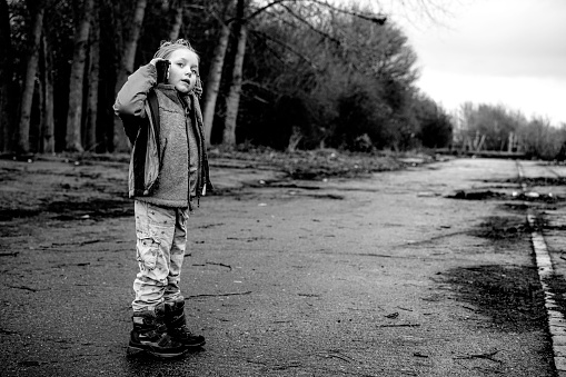 This is a black and white shot of a6year old girl in a derelict area in the road lined with trees on her cell phone during the Fall.