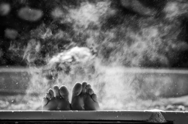 Black and white photo of a woman in a hot tub bath with her feet up and the side of the bath and steam rising above the water.