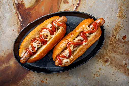 Overhead view of 2 artisan hot-dogs in a brioche bun with onions and pickles resting on a griddle pan. Colour, horizontal, some copy space against a rusty metal background.