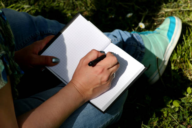 istock Woman and student sitting on grass, takes notes in notebook, learning and writes thoughts, writes book, she is preparing his final exams doing homework and thinking, writing something in his notebook. 1159603233