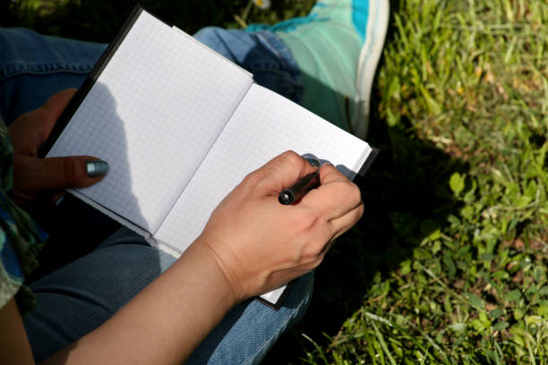 istock Woman and student sitting on grass, takes notes in notebook, learning and writes thoughts, writes book, she is preparing his final exams doing homework and thinking, writing something in his notebook. 1159603283