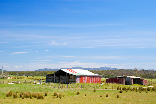 Idyllic rural landscape with green meadows, a blue sky with light streaky clouds and an old, red-painted hay shed and other outbuildings made from corrugated iron. Taken in northern Tasmania, Australia