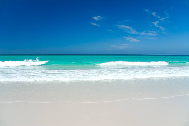 Perfect white sand beach on the remote and beautiful Bay of Fires in north-western Tasmania, Australia. The water is turquoise, and white-egded waves break cleanly on the shore. Overhead there is a deep blue sky with a few light cirrus clouds.