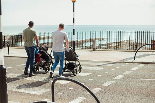 Two fathers pushing their sons in strollers across a zebra crossing in Tynemouth, UK. You can see the beach across the road.