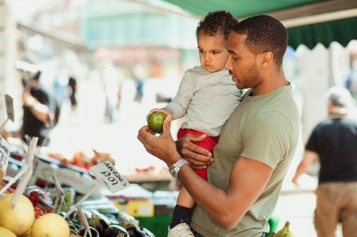 Father holding his son while they are out shopping, looking at different fruits.