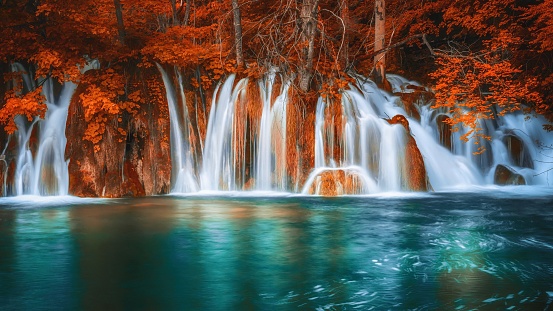 A set of waterfalls looking dreamy and ethereal, with smooth white water running into a calm pool of water below. The foliage is a vibrant red and orange, looking like a fantasy autumn season nature scene.