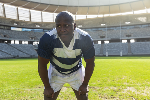 Portrait close up of tired handsome African American male rugby player standing with his hands on thighs in stadium on sunny day.