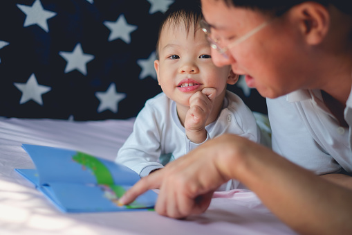 Padre y lindo pequeño asiático 2 - 3 años niño niño leyendo el libro de cuentos antes de acostarse, acostado en la cama en casa photo