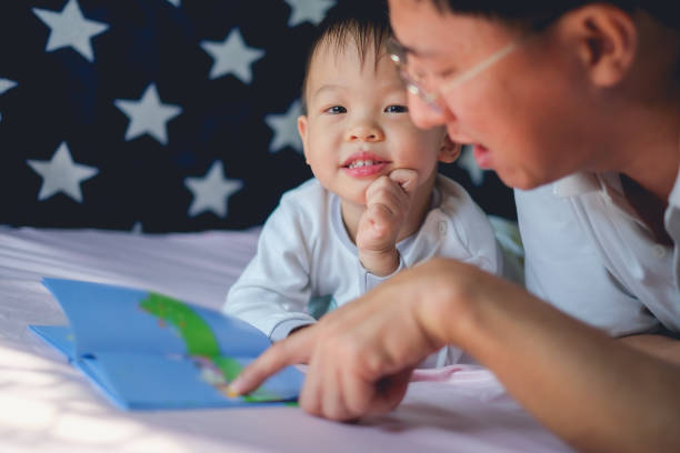 Father and Cute little Asian 2 - 3 years old toddler boy child reading bedtime story book, lying in the bed at home stock photo