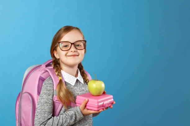 retrato de una niña colegiala en gafas con una mochila y almuerzo boxeo sobre un fondo azul. regreso a la escuela. el concepto de educación. copiar espacio - lunch lunch box child school fotografías e imágenes de stock