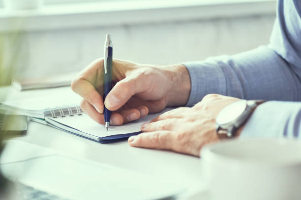 istock Businessman writing note on a notebook close-up. Business man working at office desk. Close up of empty notebook on a blackboard with office supplies. 1159657924
