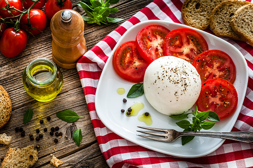 Top view of a healthy fresh burrata cheese with sliced tomatoes in a white plate placed on a gingham tablecloth shot on rustic wooden table. Ripe tomatoes, olive oil, basil, pepper and crostini complete the composition. Predominant colors are red and white. Low key DSRL studio photo taken with Canon EOS 5D Mk II and Canon EF 100mm f/2.8L Macro IS USM