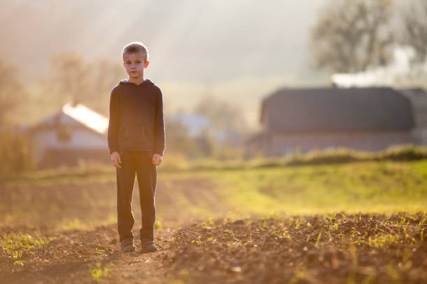 Young blond serious tired child boy standing alone on field after harvest on late summer or autumn sunny day on distant foggy blurred blue panorama of small houses among green trees background. Young blond serious tired child boy standing alone on field after harvest on late summer or autumn sunny day on distant foggy blurred blue panorama of small houses among green trees background. sad child standing stock pictures, royalty-free photos & images