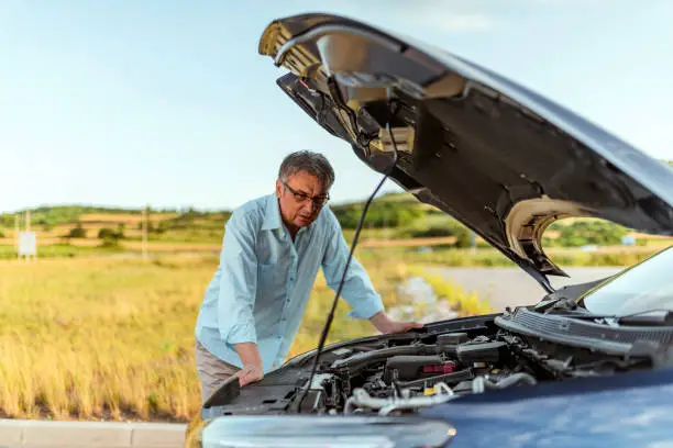 Senior Man examining a broken car on a sunny day. Photo of senior businessman looking under the hood of breakdown car at outdoor. Gray hair stressed man having trouble with his broken car looking in frustration on the failed engine.