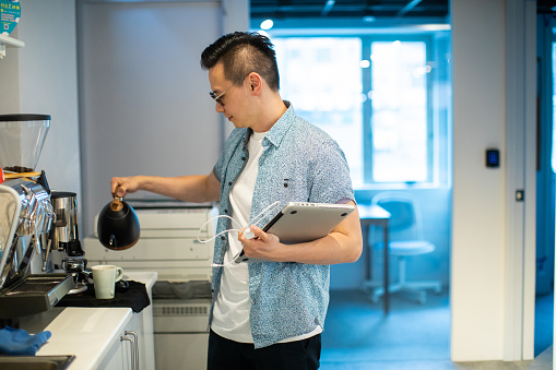 Young Asian coworker making coffee at work, using coffee maker, holding digital tablet