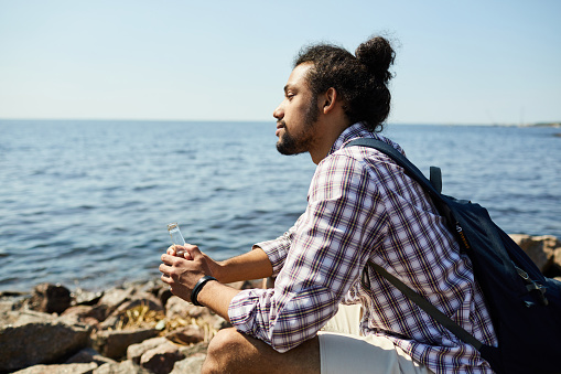 Side view portrait of contemporary Latin-American man sitting on rocks and looking at sea in Summer, copy space