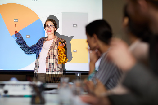 Waist up portrait of smiling businesswoman looking at audience standing against projector screen with data graph, copy space