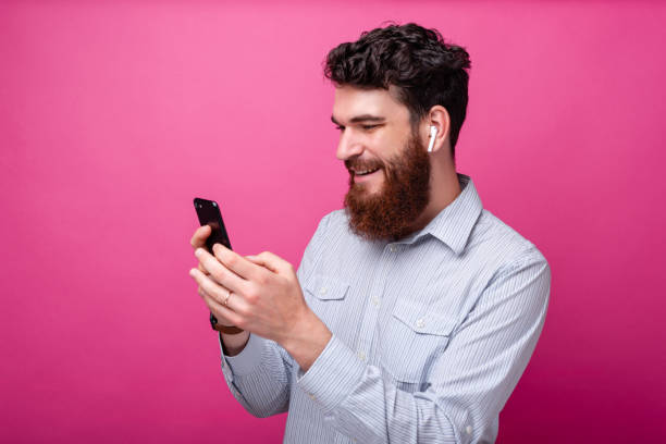 istock Young bearded man using phone and his earpods on pink background. Wireless technology. 1159675857