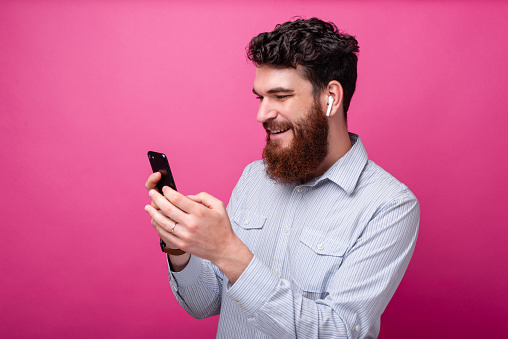 Young bearded man using phone and his earpods on pink background. Wireless technology.