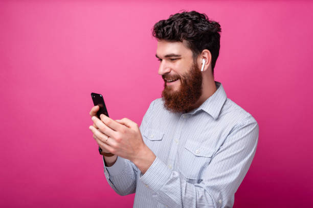 Young bearded man using phone and his earpods on pink background. Wireless technology. stock photo