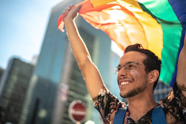 joven hombre gay sosteniendo la bandera del arco iris durante el desfile del orgullo - gay pride flag gay pride gay man homosexual fotografías e imágenes de stock