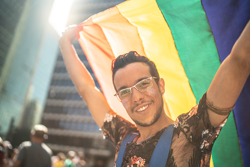 Young gay man holding rainbow flag and looking at camera during pride parade